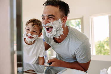 Happy father and son having fun while shaving in bathroom. Young man and little boy with shaving foam on their faces looking into the bathroom mirror and laughing. - JLPSF26347