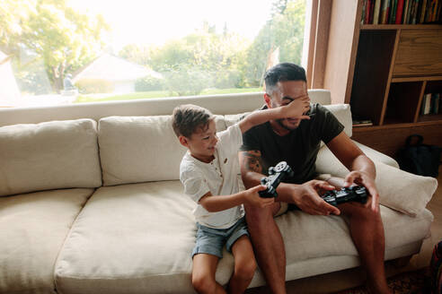 Fröhliche Familie von Vater und Sohn, die Spaß beim Spielen von Videospielen zu Hause haben. Kleiner Junge bedeckt die Augen seines Vaters beim Spielen von Videospielen im Wohnzimmer. - JLPSF26351