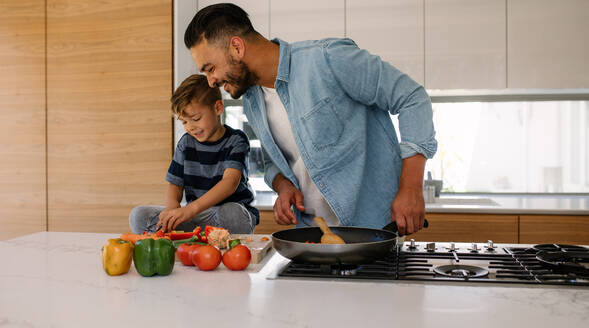 Lächelnder Vater sieht seinen kleinen Sohn an, der Gemüse auf einem Schneidebrett schneidet. Vater und Sohn kochen in der Küche. - JLPSF26356
