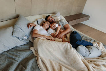 Kleiner Junge benutzt ein Mobiltelefon, während er mit seinem Vater und seiner Mutter auf dem Bett liegt. Junge Familie im Schlafzimmer, die auf ein Mobiltelefon schaut. - JLPSF26359