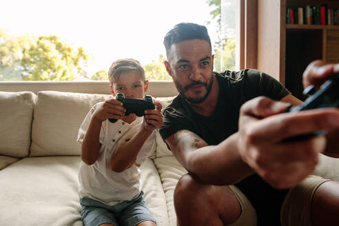 Vater und Sohn spielen ein Videospiel zu Hause. Junger Mann und kleiner Junge sitzen auf dem Sofa im Wohnzimmer und spielen ein Videospiel. - JLPSF26363