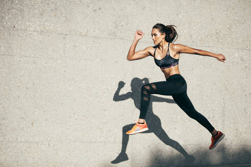 Young woman with fit body jumping and running against grey background. Female model in sportswear exercising outdoors. - JLPSF26393