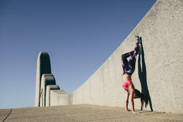 Physically fit woman doing hand stand outdoors. Healthy female standing on hands with feet lifted up against a wall. - JLPSF26422