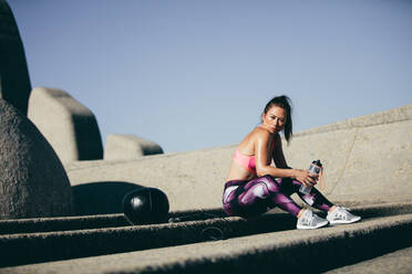 Fitness-Frau, die mit einer Wasserflasche im Freien sitzt. Sportlerin, die nach einer Trainingseinheit eine Pause einlegt. - JLPSF26423