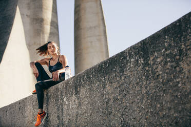 Sporty woman sitting outdoors after fitness training. Healthy female in sportswear taking a break after workout. - JLPSF26439