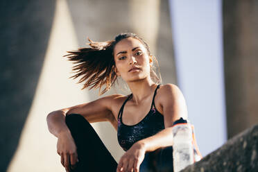 Fitte junge Frau sitzt nach einem morgendlichen Workout im Freien. Weibliches Fitnessmodel entspannt sich und starrt in die Kamera. - JLPSF26440