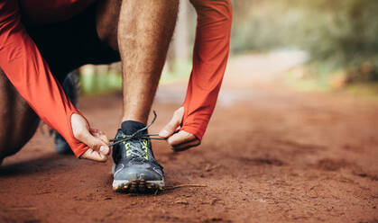 Close up shot of an athlete tying his shoe laces. Man tightening his shoe laces resting on one knee on a mud track. - JLPSF26445