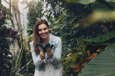 Happy woman standing in the plant nursery wearing hand gloves. Smiling female in the garden center looking at camera. - JLPSF26506