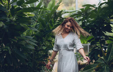 Lächelndes junges weibliches Modell, das in einem Gartencenter spazieren geht. Glückliche junge Frau, die sich im Gewächshaus vergnügt. - JLPSF26509