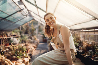 Portrait of smiling woman sitting in greenhouse. Beautiful gardener smiling at plant nursery. - JLPSF26537