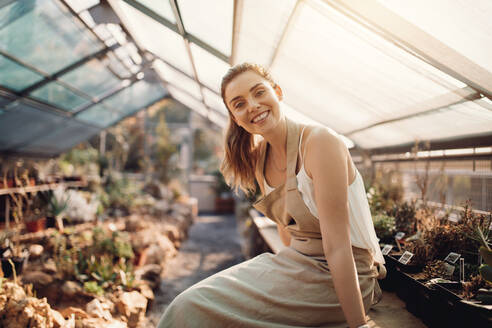 Porträt einer lächelnden Frau, die in einem Gewächshaus sitzt. Schöne Gärtnerin, die in einer Gärtnerei lächelt. - JLPSF26537