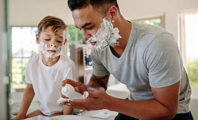 Father and son applying shaving foam on their faces in bathroom. Man and little boy putting shaving foam on their faces while shaving in bathroom. - JLPSF26547