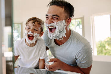 Man and little boy with shaving foam on their faces looking into the bathroom mirror and laughing. Father and son having fun while shaving in bathroom. - JLPSF26548