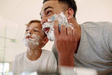 Father and son making funny faces while shaving in bathroom. Young man and little boy with shaving foam on their faces are shaving and looking into the mirror. - JLPSF26549