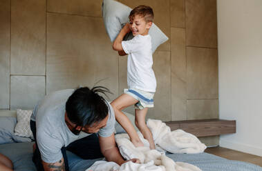 Kleiner Junge schlägt einen Mann mit einem Kissen auf dem Bett. Vater und Sohn genießen es, eine Kissenschlacht im Schlafzimmer zu spielen. - JLPSF26552