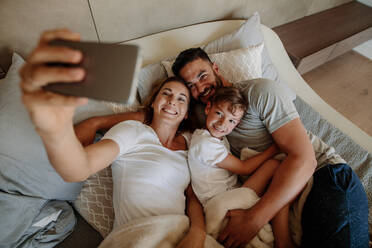 Glückliche junge Frau, die ein Selfie mit ihrer Familie macht. Die Familie liegt auf dem Bett und macht ein Selfie zu Hause. - JLPSF26553