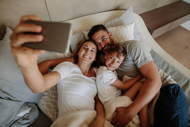 Happy young woman taking selfie with her family. Family lying on bed and taking selfie at home. - JLPSF26553