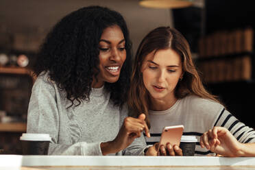 Zwei Frauen sitzen in einem Restaurant und schauen auf ein Handy und unterhalten sich. Freunde sitzen in einem Café mit Kaffee und Snacks auf dem Tisch und schauen auf ein Mobiltelefon. - JLPSF26555