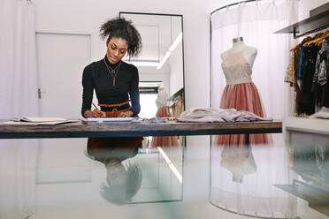 Modedesignerin, die an ihrem Tisch sitzend einen Entwurf skizziert. Modeunternehmerin, die an ihrem Schreibtisch in ihrem Stoffgeschäft in eine Zeichnung vertieft ist. - JLPSF26558