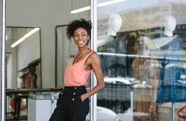 Lächelnde Kleidungsdesignerin in ihrem Stoffladen mit ausgestellter Designerkleidung. Unternehmerin steht am Eingang ihres Modestudios. - JLPSF26566