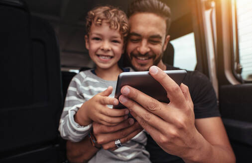 Aufnahme eines kleinen Jungen mit seinem Vater, der auf einer Autoreise ein digitales Tablet benutzt. Glücklicher Vater und Sohn sitzen hinten im Auto mit einem digitalen Tablet. - JLPSF26568