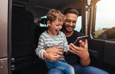 Aufnahme eines glücklichen Vaters und seines Sohnes, die hinten im Auto sitzen und auf ein digitales Tablet schauen. Junger Mann und kleiner Junge benutzen ein digitales Tablet während einer Autoreise. - JLPSF26569