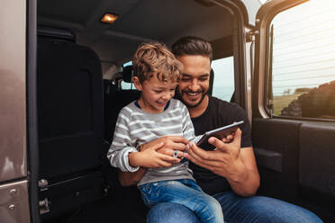 Vater und Sohn sitzen hinten im Auto und lächeln, während sie ein digitales Tablet benutzen. Junger Mann mit kleinem Jungen, der ein digitales Tablet auf einer Autoreise hält. - JLPSF26570