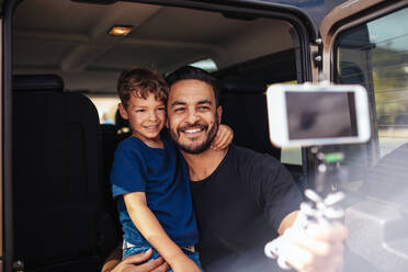Hübscher junger Vater und sein süßer kleiner Sohn sitzen auf der Rückbank des Autos und machen ein Selfie mit einem Smartphone. Vater und Sohn auf einer Autoreise machen ein Selfie. - JLPSF26573