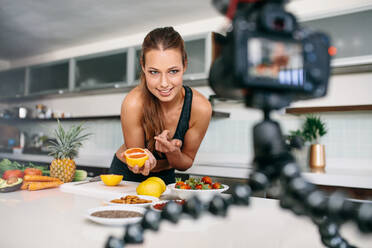 Junge Frau, die mit der Kamera lebensmittelbasierte Videoinhalte aufnimmt. Die Frau zeigt auf eine aufgeschnittene Orange, die in die Kamera zeigt. - JLPSF26581