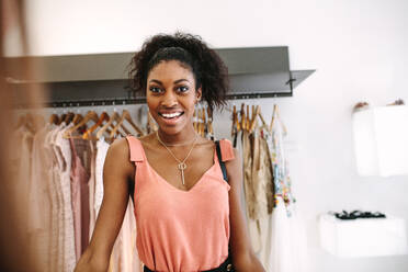 Lächelnde Frau, die in einem Bekleidungsgeschäft mit Designer-Kleidung im Hintergrund steht. Zufriedene Kundin in einem Bekleidungsgeschäft. - JLPSF26598