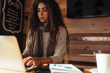 Woman sitting at a restaurant table working on laptop. Freelancer sitting in cafe with a coffee, notepad and pen working on laptop computer. - JLPSF26641