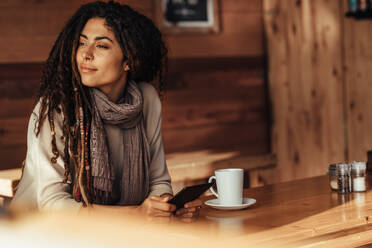 Frau sitzt in einem Restaurant und hält ihr Handy in der Hand und schaut weg. Frau sitzt in einem Café mit Kaffee auf dem Tisch. - JLPSF26650
