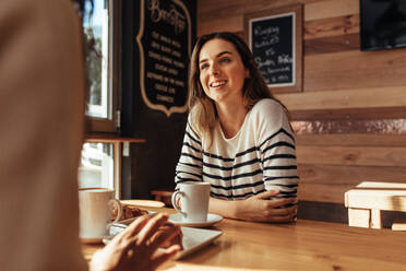 Smiling woman sitting in a restaurant talking to her friend. Friends sitting at a cafe with coffee and snacks on the table. - JLPSF26652