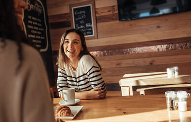 Lächelnde Frau, die in einem Restaurant sitzt und sich mit ihrer Freundin unterhält. Freunde sitzen in einem Café mit Kaffee und Snacks auf dem Tisch. - JLPSF26653