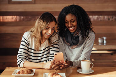 Zwei lächelnde Frauen sitzen in einem Restaurant und schauen auf ihr Handy. Freunde sitzen in einem Café mit Kaffee und Snacks auf dem Tisch und schauen auf ihr Handy. - JLPSF26656