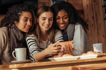 Drei lächelnde Frauen sitzen in einem Restaurant und schauen auf ein Handy. Freunde sitzen in einem Café mit Kaffee und Snacks auf dem Tisch und schauen auf ein Handy. - JLPSF26657