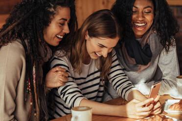 Drei Frauen sitzen in einem Restaurant und schauen auf ihr Handy und lachen. Freunde sitzen in einem Café mit Kaffee und Snacks auf dem Tisch und schauen auf ein Handy. - JLPSF26658