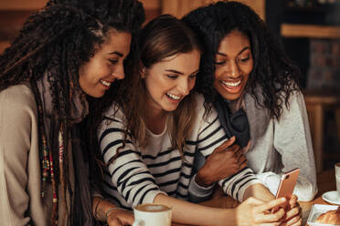 Drei Frauen sitzen in einem Restaurant und schauen auf ihr Handy und lachen. Freunde sitzen in einem Café mit Kaffee und Snacks auf dem Tisch und schauen auf ein Handy. - JLPSF26659