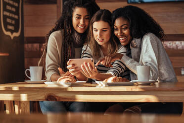 Drei Frauen sitzen in einem Restaurant und schauen auf ihr Handy und lachen. Freunde sitzen in einem Café mit Kaffee und Snacks auf dem Tisch und schauen auf ein Handy. - JLPSF26660