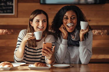 Zwei Frauen trinken Kaffee und lächeln mit einem Milchschnurrbart in einem Café. Freunde sitzen in einem Café mit Kaffee und Snacks auf dem Tisch und halten ein Mobiltelefon. - JLPSF26661