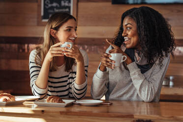 Two women drinking coffee and smiling at each other pointing towards milk mustache in coffee shop. Friends sitting at a cafe with coffee and snacks on the table. - JLPSF26662