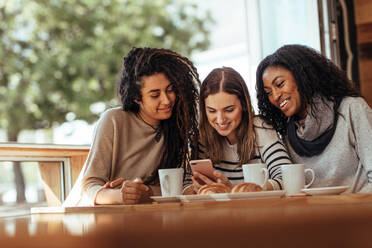 Drei Frauen sitzen in einem Restaurant und schauen lächelnd auf ihr Handy. Freunde sitzen in einem Café mit Kaffee und Snacks auf dem Tisch und schauen auf ein Handy. - JLPSF26665