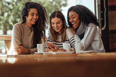 Drei Frauen sitzen in einem Restaurant und schauen auf ihr Handy und lachen. Freunde sitzen in einem Café mit Kaffee und Snacks auf dem Tisch und schauen auf ein Handy. - JLPSF26666