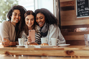Drei Frauen sitzen in einem Restaurant und schauen lächelnd auf ihr Handy. Freunde sitzen in einem Café mit Kaffee und Snacks auf dem Tisch und schauen auf ein Handy. - JLPSF26667