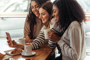 Drei Frauen sitzen in einem Restaurant und schauen lächelnd auf ihr Handy. Freunde sitzen in einem Café mit Kaffee und Snacks auf dem Tisch und schauen auf ein Handy. - JLPSF26668