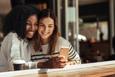 Zwei lächelnde Frauen sitzen in einem Restaurant und machen ein Selfie mit dem Handy. Freunde sitzen in einem Café mit Kaffee und Snacks auf dem Tisch und schauen auf ein Handy. - JLPSF26670