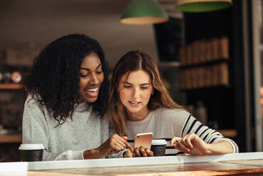 Zwei Frauen sitzen in einem Restaurant und schauen auf ein Handy und unterhalten sich. Freunde sitzen in einem Café mit Kaffee und Snacks auf dem Tisch und schauen auf ein Mobiltelefon. - JLPSF26671