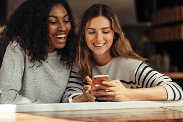 Two smiling women sitting in a restaurant looking at mobile phone and laughing. Friends sitting at a cafe smiling looking at a mobile phone. - JLPSF26672