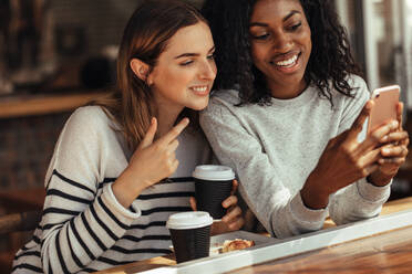 Two women sitting in a restaurant looking at mobile phone and talking. Friends sitting at a cafe with coffee and snacks on the table taking selfie using a mobile phone. - JLPSF26673