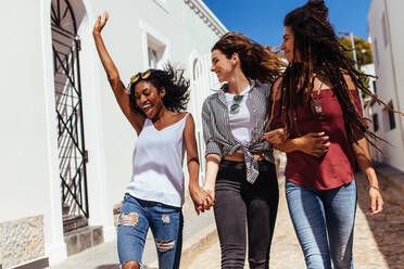 Three women wearing fashionable clothes walking on street on a sunny day. Young woman jumping in joy while walking with friends on a street. - JLPSF26675